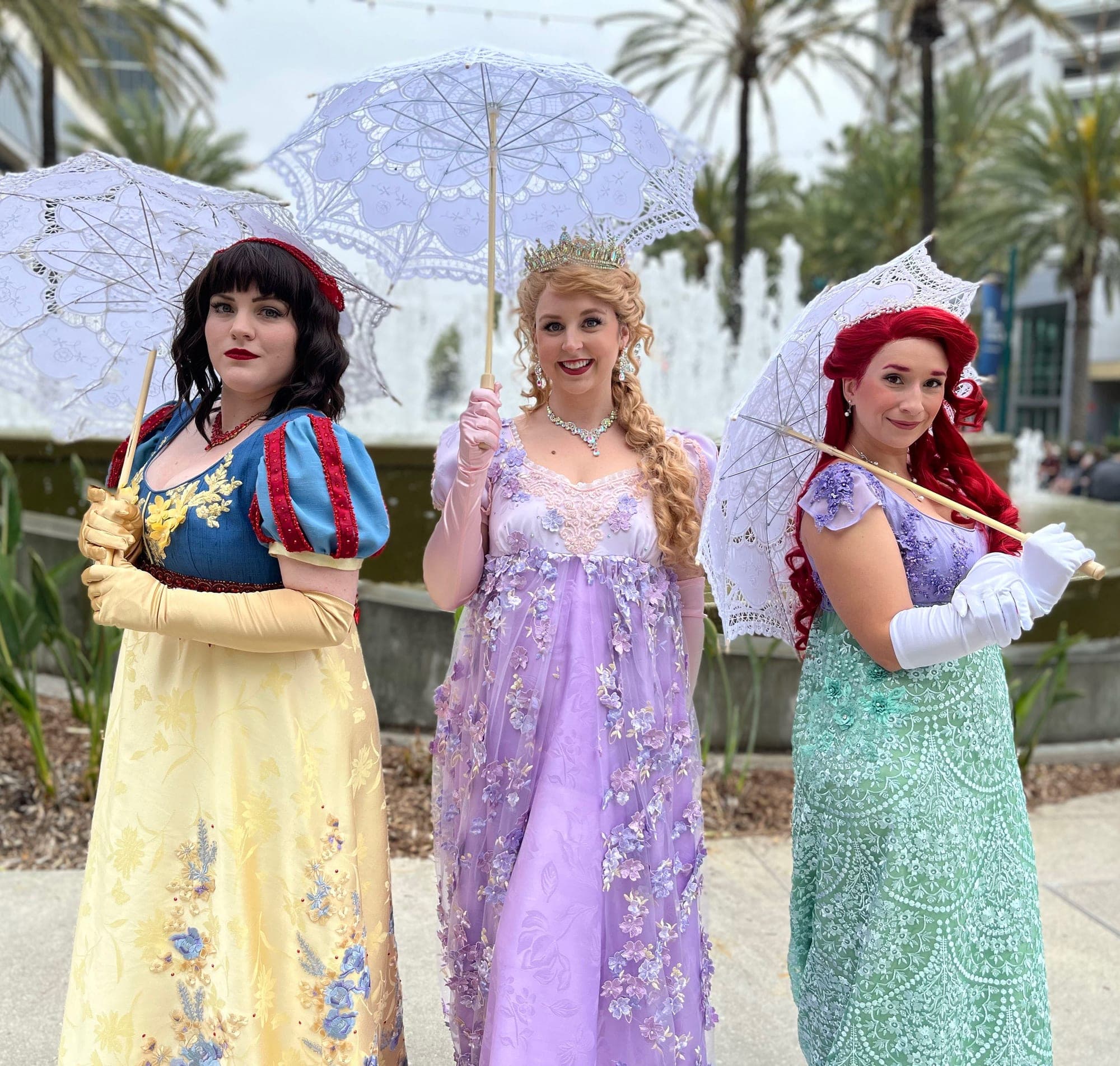 Three princess cosplayers with lace parasols — Snow White, Rapunzel, and Ariel