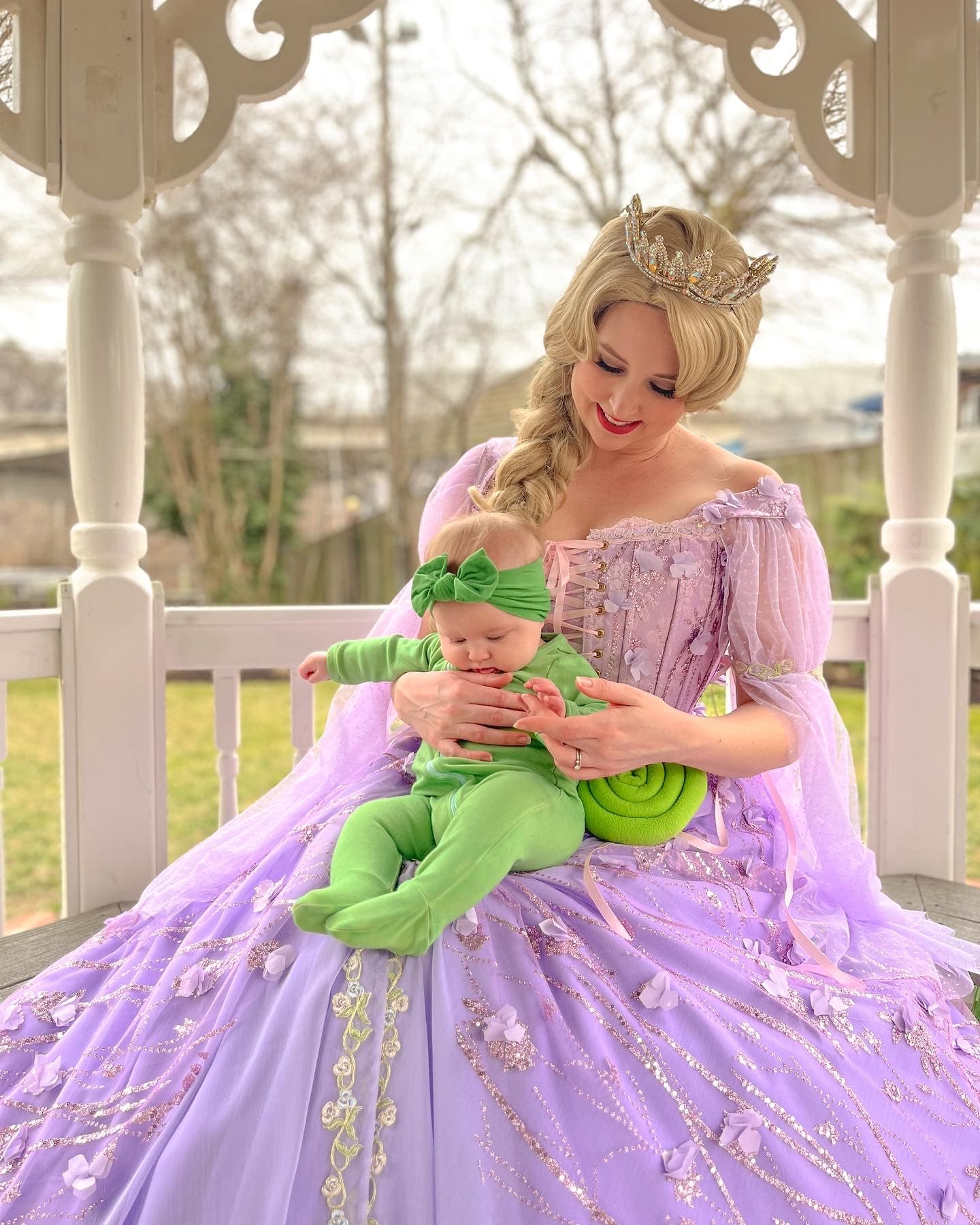 Woman in a lavender Rapunzel gown holding a baby in green on a gazebo